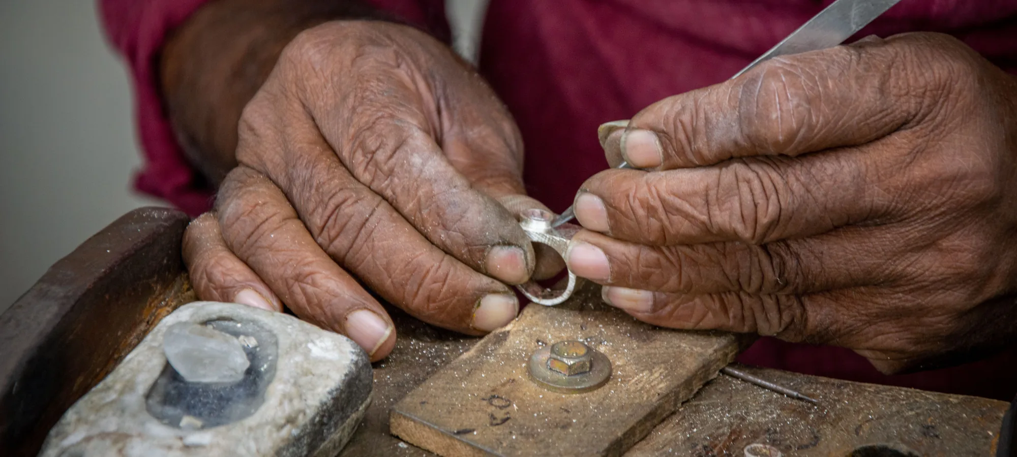 A craftsman’s weathered hands holding a small silver ring and a pointed tool over a wooden bench block